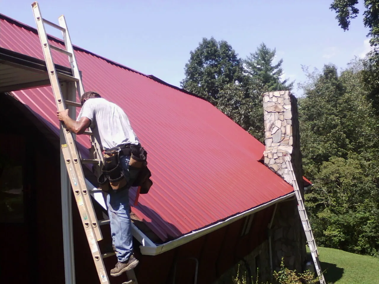 Roofer on a ladder beside a red metal roof and stone chimney — Soffit & Fascia Repair by Roofing Brothers in Hailey